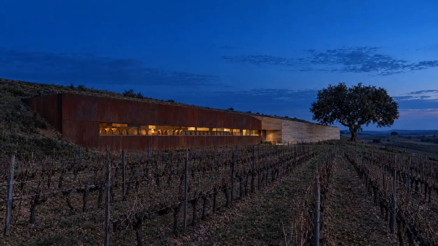 Bodega de acero corten y hormigón semienterrada entre viñedos, con una ventana corrida encendida a la hora azul